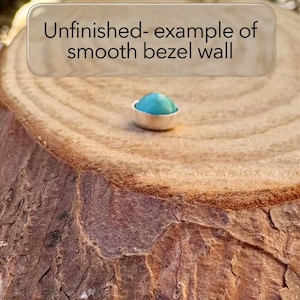 May include: A small, silver bezel setting with a smooth, turquoise-colored stone. The setting is displayed on a textured, brown wooden surface. Text above reads "Unfinished- example of smooth bezel wall."