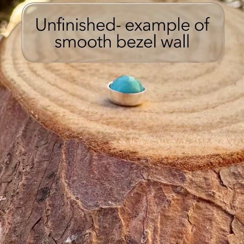 May include: A small, silver bezel setting with a smooth, turquoise-colored stone. The setting is displayed on a textured, brown wooden surface. Text above reads "Unfinished- example of smooth bezel wall."