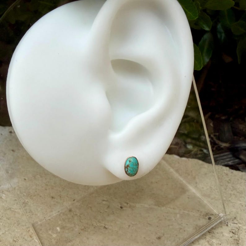 May include: A single turquoise stud earring with a silver-colored bezel is displayed on a white ear model. The oval-shaped stone has a mottled blue-green color. The earring is set against a blurred background of greenery.