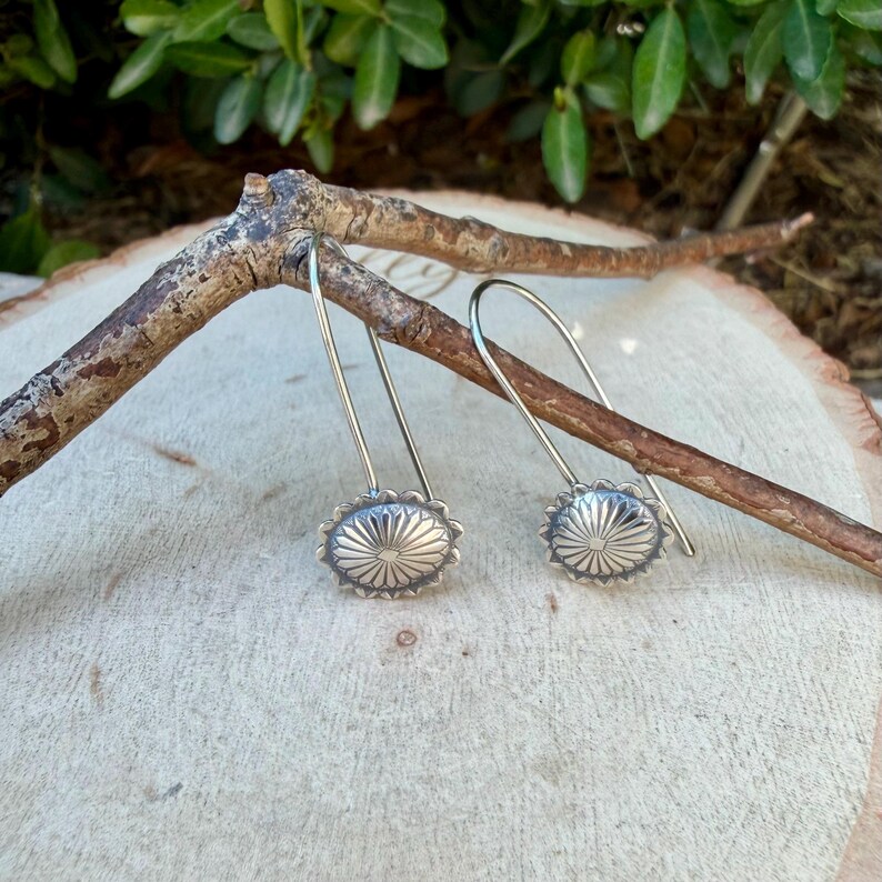 May include: A pair of silver drop earrings featuring circular, sunburst-patterned charms with oxidized detailing. Each earring has a long, curved wire hook that extends upward, showcasing a radial design with fine, etched lines radiating from the center.
