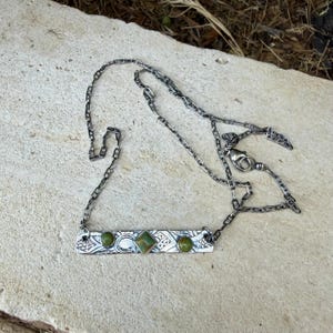 May include: A silver-toned metal necklace featuring a rectangular bar pendant with an etched pattern. The pendant is set with two round green stones on the ends and a central square green stone. The chain is a flat, interlocking link style.