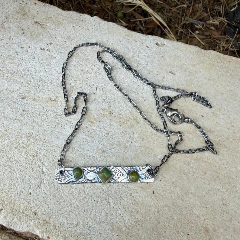 May include: A silver-toned metal necklace featuring a rectangular bar pendant with an etched pattern. The pendant is set with two round green stones on the ends and a central square green stone. The chain is a flat, interlocking link style.
