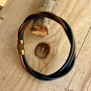 May include: A multi-strand bracelet composed of dark brown and black leather cords twisted together. The bracelet features a cylindrical brass-toned metal magnetic clasp that secures the ends of the leather cords into a circular shape.
