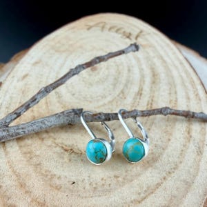 May include: Silver leverback earrings featuring round, turquoise stones. The earrings are presented on a wooden surface with a small branch, highlighting their natural style. The turquoise stones have brown veining.