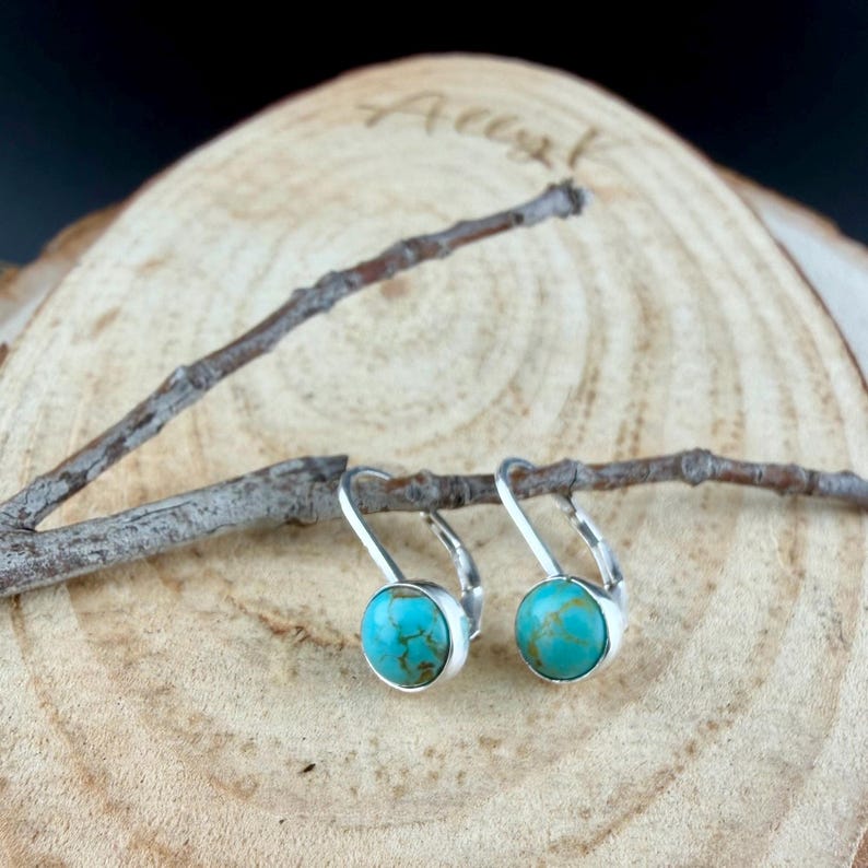 May include: Silver leverback earrings featuring round, turquoise stones. The earrings are presented on a wooden surface with a small branch, highlighting their natural style. The turquoise stones have brown veining.