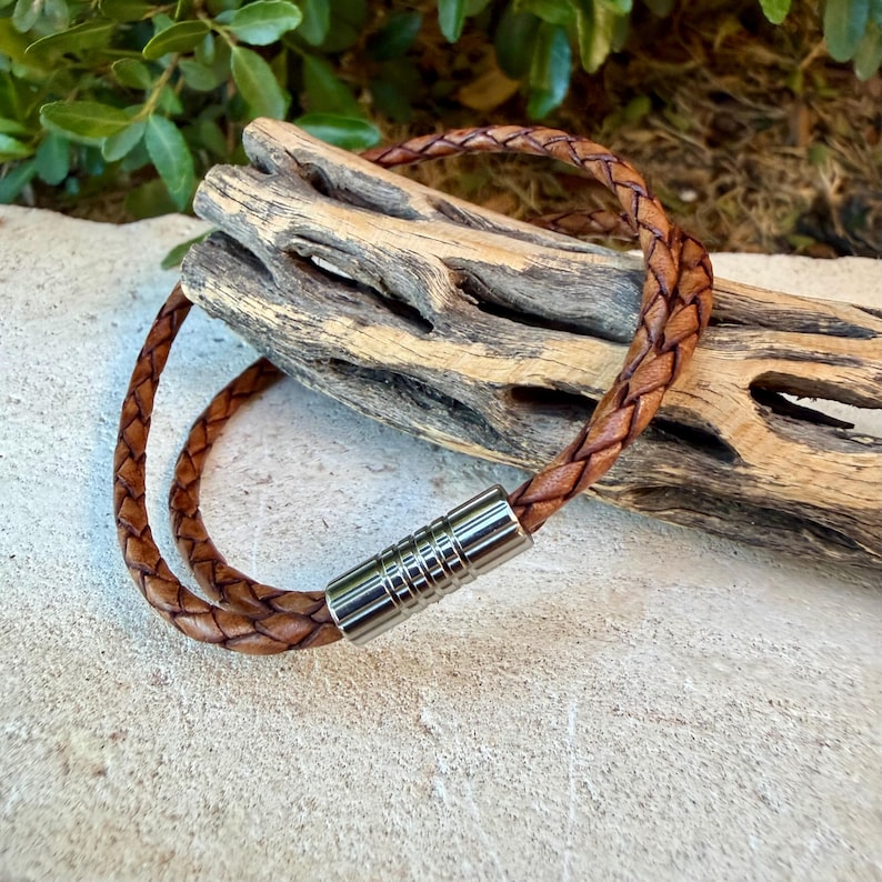 May include: A brown braided leather bracelet featuring a cylindrical silver-toned metal clasp with a ribbed texture. The bracelet is formed from a single strand of woven leather that loops around to connect at the metallic fastener.