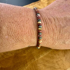 May include: A beaded bracelet featuring a variety of earth-toned beads, including shades of brown, beige, green, and red. The bracelet is strung on a flexible cord and is approximately 7 inches in circumference.
