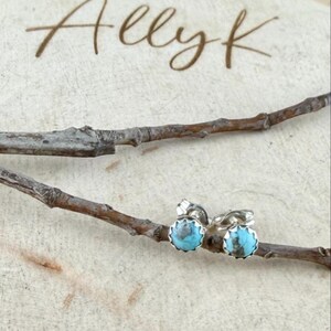 May include: A pair of turquoise stud earrings with a silver setting. The earrings are round with a scalloped edge. The image also includes the text "Allyk" in a handwritten font, and is set against a light wood background with a twig.