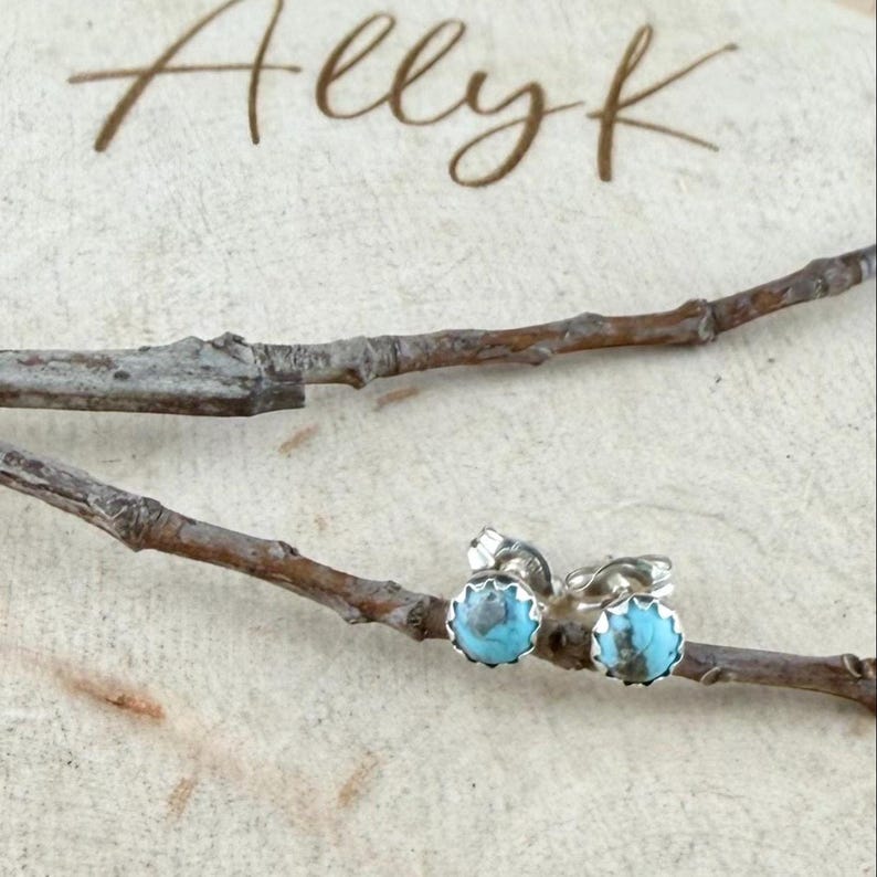 May include: A pair of turquoise stud earrings with a silver setting. The earrings are round with a scalloped edge. The image also includes the text "Allyk" in a handwritten font, and is set against a light wood background with a twig.