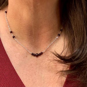 May include: A delicate silver necklace featuring small, dark red beads. The beads are evenly spaced along the chain, with a cluster of larger beads at the center. The necklace is shown against a skin tone background, highlighting the jewelry's design.