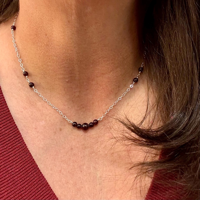 May include: A delicate silver necklace featuring small, dark red beads. The beads are evenly spaced along the chain, with a cluster of larger beads at the center. The necklace is shown against a skin tone background, highlighting the jewelry's design.