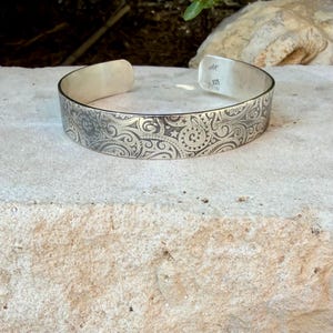 May include: A silver cuff bracelet featuring an intricate engraved pattern of swirling lines, spirals, and small stars. The interior of the band is stamped with the letters AK and the number 925, indicating sterling silver composition.