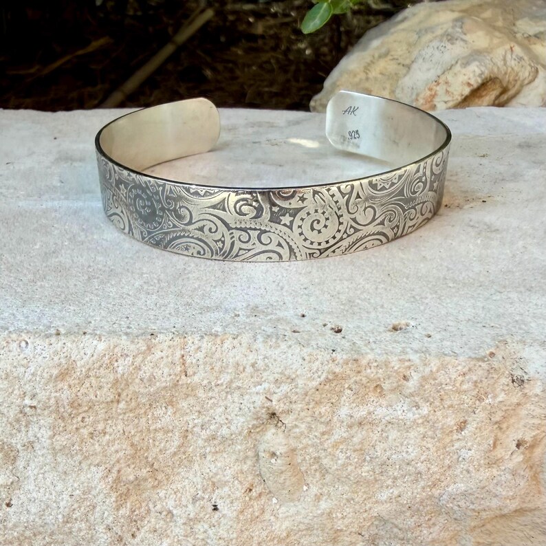 May include: A silver cuff bracelet featuring an intricate engraved pattern of swirling lines, spirals, and small stars. The interior of the band is stamped with the letters AK and the number 925, indicating sterling silver composition.