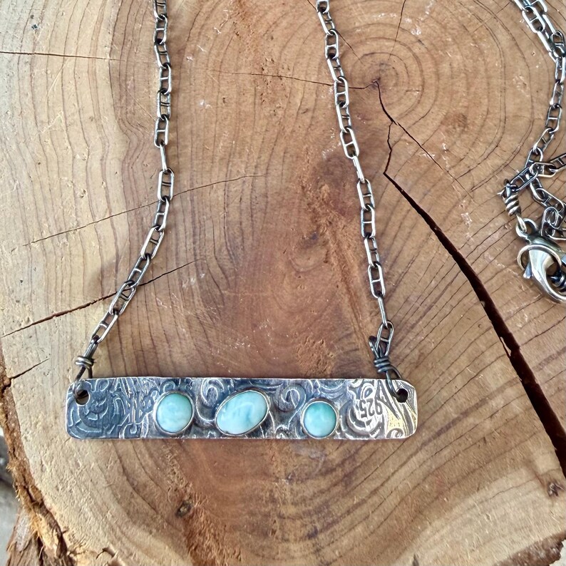 May include: A sterling silver necklace featuring a rectangular bar pendant with an etched scroll pattern and three oval light blue larimar stones. The pendant is attached to a silver chain with a lobster clasp and a 925 hallmark stamp.