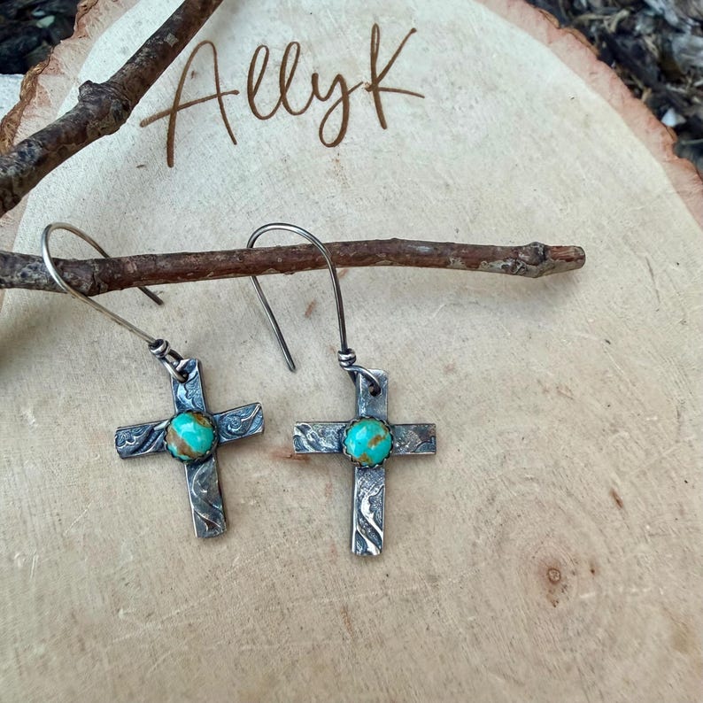 May include: A pair of silver cross earrings with turquoise-colored centers. The crosses have an etched design and are displayed on a wooden surface. The word "Allyk" is written in cursive.