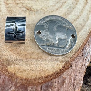 Puede incluir: Un anillo plateado con una superficie texturizada y un dise&ntilde;o de cruz, junto a una moneda plateada con una imagen de b&uacute;falo. La moneda tiene dos agujeros y se ven las inscripciones "UNITED STATES OF AMERICA" y "FIVE CENTS".
