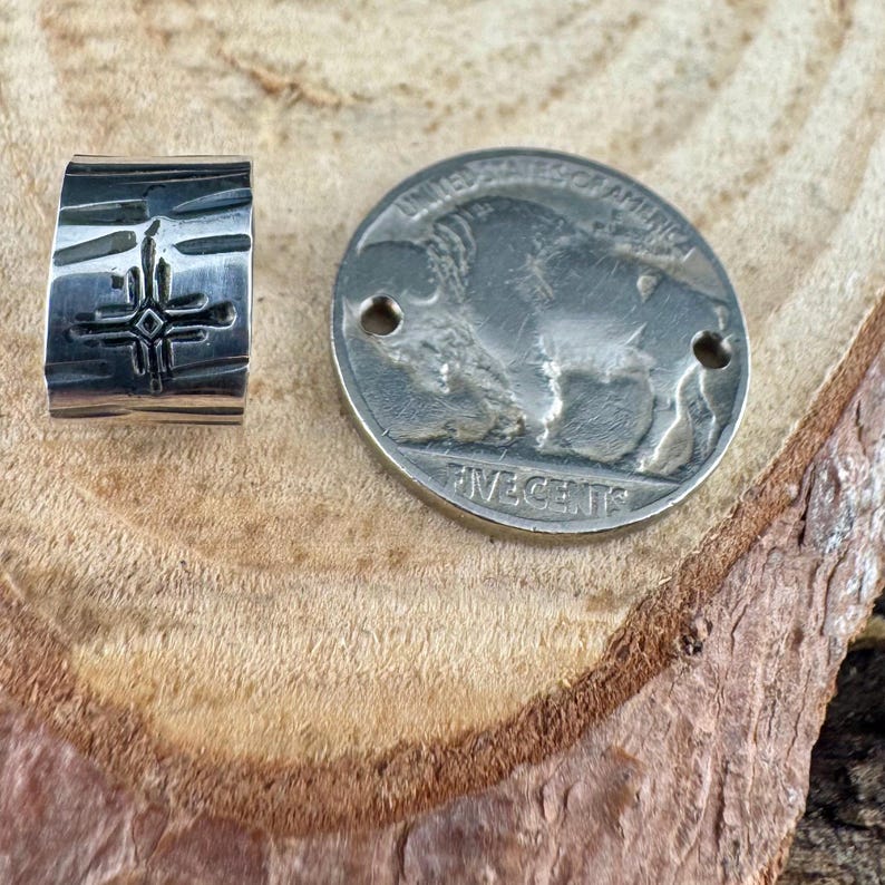 Puede incluir: Un anillo plateado con una superficie texturizada y un dise&ntilde;o de cruz, junto a una moneda plateada con una imagen de b&uacute;falo. La moneda tiene dos agujeros y se ven las inscripciones "UNITED STATES OF AMERICA" y "FIVE CENTS".