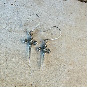 May include: A pair of silver-toned drop earrings featuring a cross-shaped design with a pointed, dagger-like base. Each earring is suspended from a curved wire hook, showcasing intricate metalwork details and a polished finish throughout the piece.