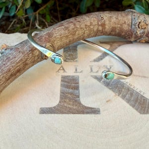 May include: A silver cuff bracelet featuring two small, oval-shaped turquoise stones set at each open end. The metal band is smooth and polished, with the turquoise stones displaying natural blue-green colour variations and dark matrix patterns.