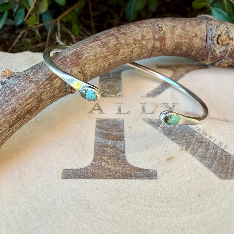 May include: A silver cuff bracelet featuring two small, oval-shaped turquoise stones set at each open end. The metal band is smooth and polished, with the turquoise stones displaying natural blue-green colour variations and dark matrix patterns.