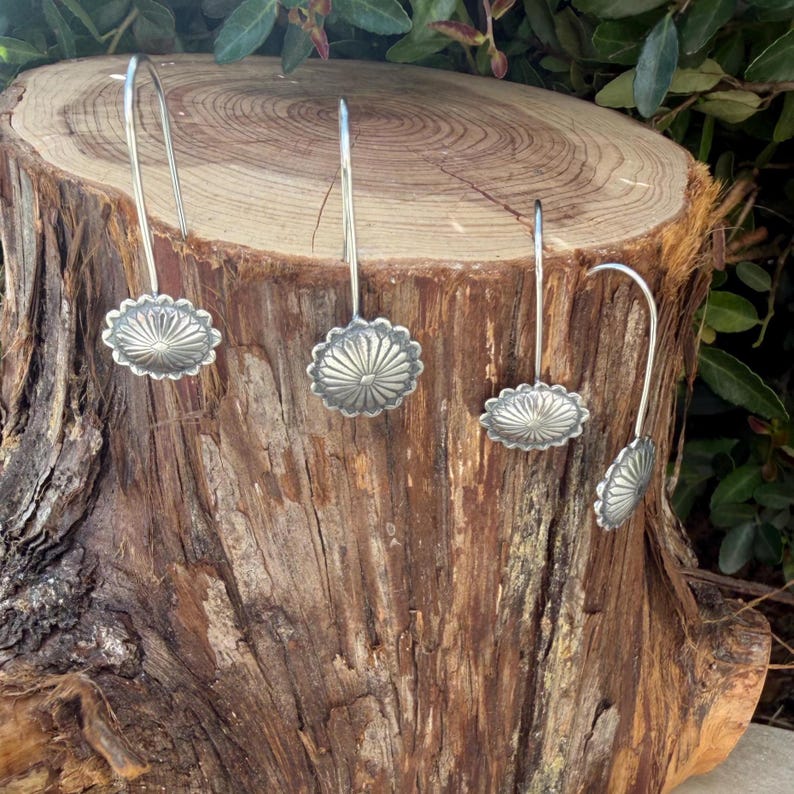 May include: Four silver-toned earrings featuring a circular, sunburst-patterned concho design. Each earring has a long, curved hook wire for suspension. The metallic pieces display a polished finish with radiating lines centered on each disc.