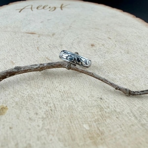 May include: A silver ring with an ornate floral design. The ring is displayed on a small branch against a light brown wooden background. The ring's design features swirling patterns and is likely made of sterling silver.