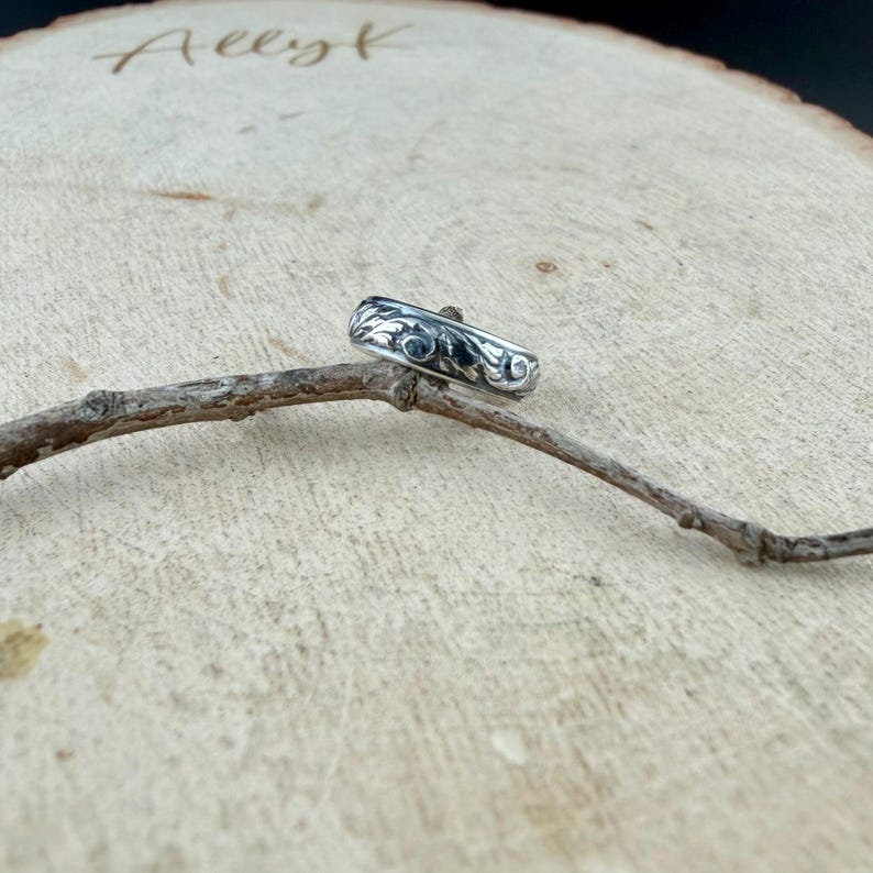 May include: A silver ring with an ornate floral design. The ring is displayed on a small branch against a light brown wooden background. The ring's design features swirling patterns and is likely made of sterling silver.
