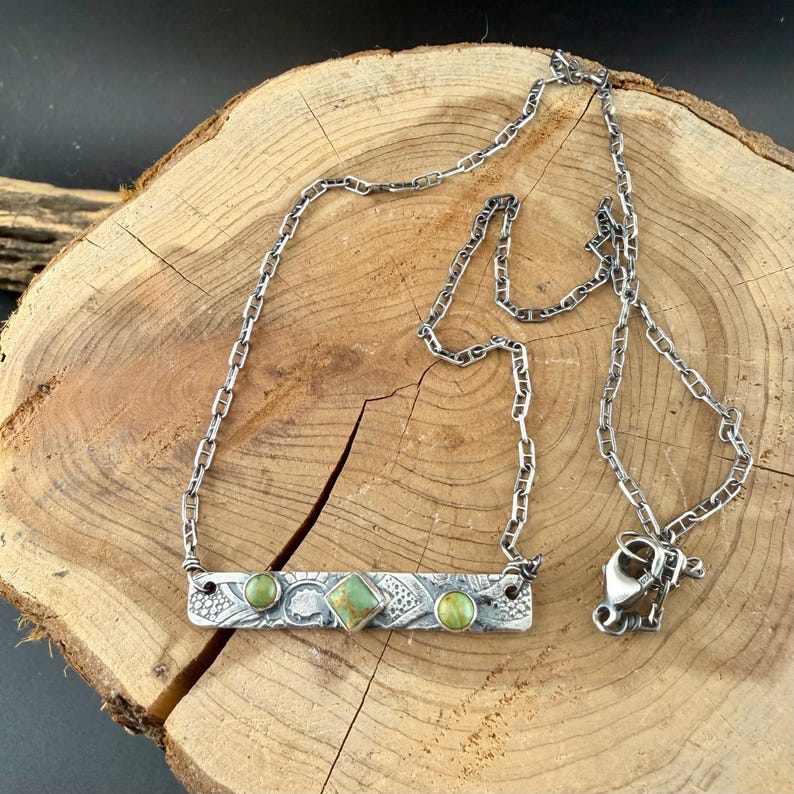 May include: A silver-toned metal necklace featuring a rectangular bar pendant with an embossed texture. The pendant is set with two small round green stones and one central square green stone. It includes a lobster claw clasp.