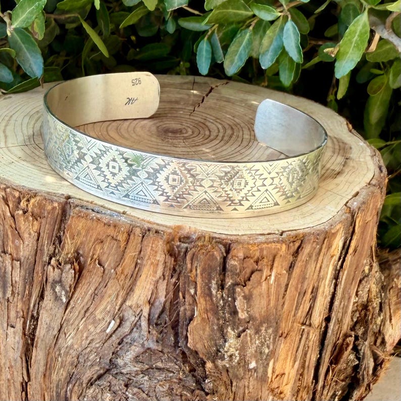 May include: A silver cuff bracelet featuring an intricate engraved geometric pattern of triangles and diamonds. The interior of the band is stamped with the text 925 and the letters AK, indicating the metal composition and a maker's mark.