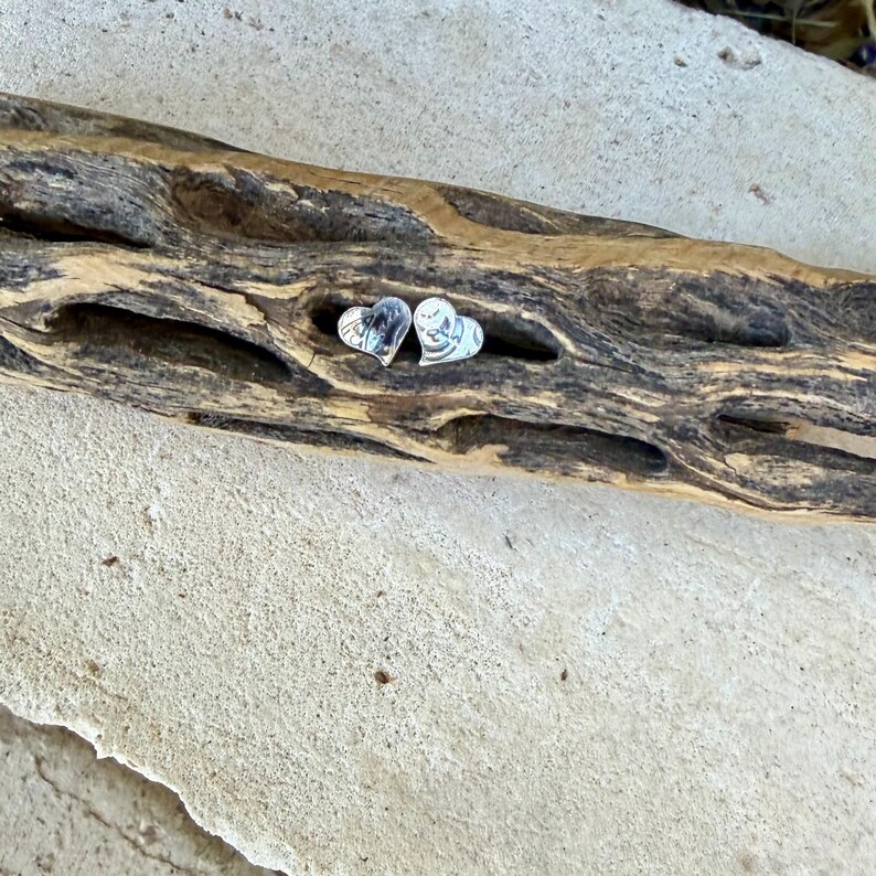 May include: A pair of small, silver-colored heart-shaped stud earrings. Each earring features a smooth, reflective surface with a subtle, etched line detail that creates a stylized, abstract pattern across the metal heart shape.