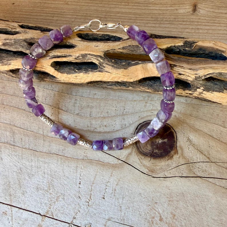 Puede incluir: Una pulsera que presenta cuentas c&uacute;bicas de amatista morada facetada dispuestas en un patr&oacute;n circular, intercaladas con peque&ntilde;as cuentas espaciadoras de metal plateado y un cierre de mosquet&oacute;n plateado para una pieza de joyer&iacute;a uniforme.