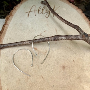 Puede incluir: Un par de pendientes de aro plateados con un dise&ntilde;o curvo y abierto con peque&ntilde;os extremos esf&eacute;ricos. Los pendientes est&aacute;n colocados cerca de una rama de madera marr&oacute;n sobre una superficie de madera clara con el texto grabado Ally K.