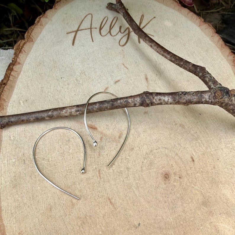 Puede incluir: Un par de pendientes de aro plateados con un dise&ntilde;o curvo y abierto con peque&ntilde;os extremos esf&eacute;ricos. Los pendientes est&aacute;n colocados cerca de una rama de madera marr&oacute;n sobre una superficie de madera clara con el texto grabado Ally K.