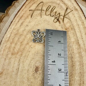 May include: A small, silver-colored butterfly charm with intricate openwork design. The charm is approximately 0.5 inches in width and is placed next to a ruler for size comparison. The background is a wooden surface with the handwritten text "Allyk".