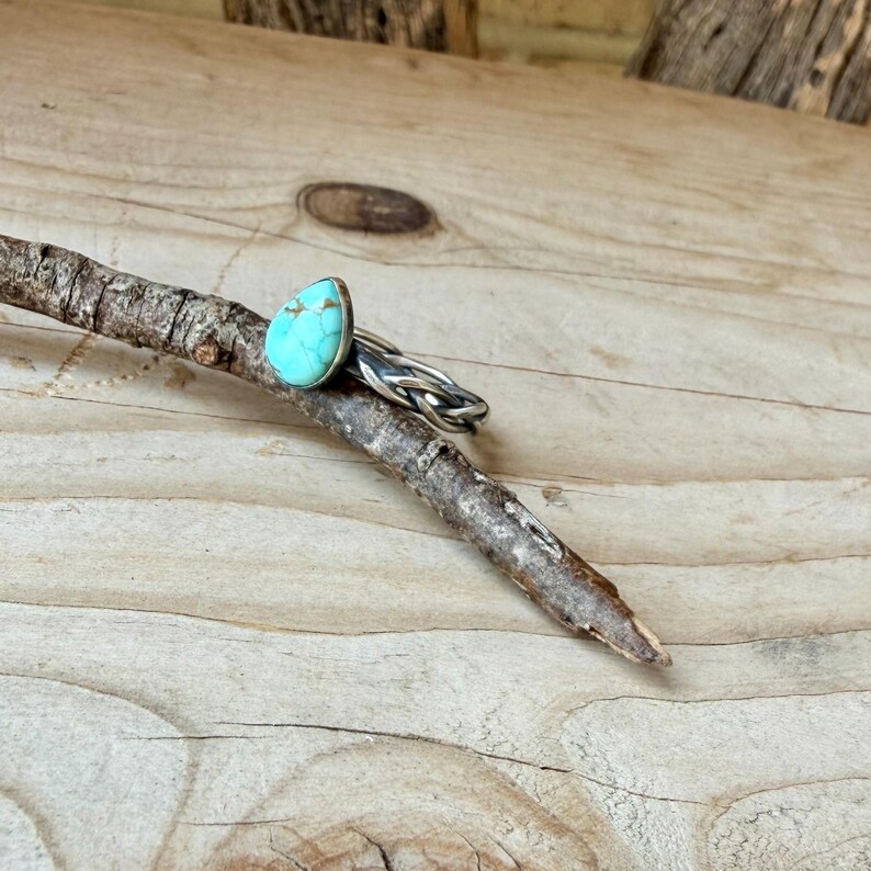 Puede incluir: Un anillo con una piedra de turquesa en forma de l&aacute;grima con patrones de matriz marr&oacute;n, engastada en un bisel de plata. La banda est&aacute; hecha de alambre de plata trenzado, creando una apariencia de trenza con un acabado met&aacute;lico texturizado.