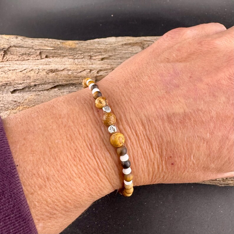May include: A beaded bracelet featuring a mix of brown, tan, white, and silver beads. The bracelet is made of various sizes and shapes of beads, including round and cylindrical ones. The bracelet is worn on a wrist.