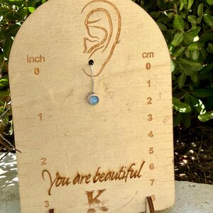May include: A silver belly button ring with a round, iridescent moonstone gem. The jewelry is displayed on a light wood board featuring an ear outline, inch and centimeter markings, and the engraved text "You are beautiful" and "K".
