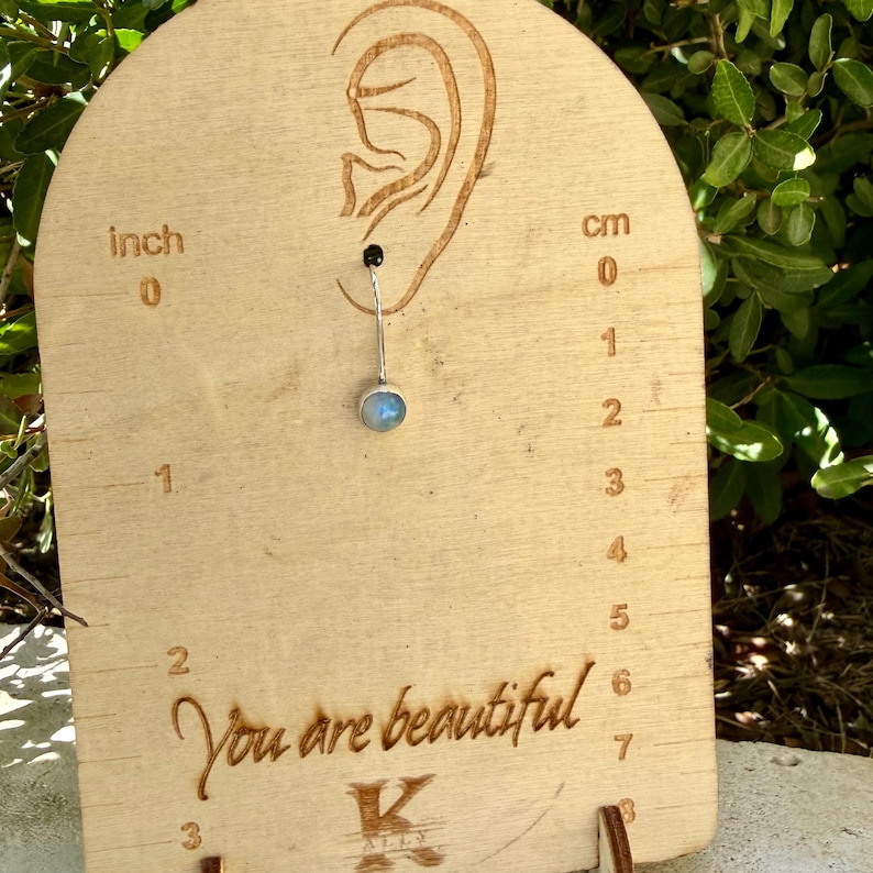 May include: A silver belly button ring with a round, iridescent moonstone gem. The jewelry is displayed on a light wood board featuring an ear outline, inch and centimeter markings, and the engraved text "You are beautiful" and "K".
