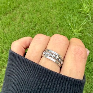 May include: A hand wearing three silver rings. The top ring has a row of clear stones. The middle ring has a row of clear stones. The bottom ring has a textured design. The background is green grass.