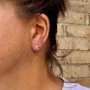 May include: A small, round, silver-toned stud earring featuring a textured, embossed surface design. The earring is worn in the earlobe, positioned below a tiny, simple silver-toned stud earring in the upper lobe area.