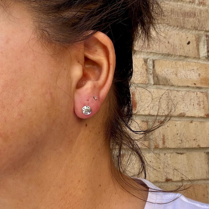 May include: A small, round, silver-toned stud earring featuring a textured, embossed surface design. The earring is worn in the earlobe, positioned below a tiny, simple silver-toned stud earring in the upper lobe area.