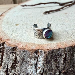 May include: A silver-toned adjustable ring with an engraved floral pattern. The ring features a round, iridescent gemstone with shades of purple, blue, and green. The ring is displayed on a wooden surface.