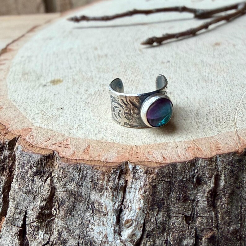 May include: A silver-toned adjustable ring with an engraved floral pattern. The ring features a round, iridescent gemstone with shades of purple, blue, and green. The ring is displayed on a wooden surface.