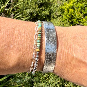 May include: A silver cuff bracelet featuring an intricate engraved swirling pattern alongside a layered bracelet consisting of a silver chain and a strand of small, round, multi-colored turquoise and earth-toned stone beads.