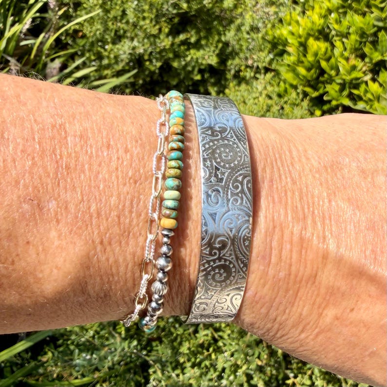 May include: A silver cuff bracelet featuring an intricate engraved swirling pattern alongside a layered bracelet consisting of a silver chain and a strand of small, round, multi-colored turquoise and earth-toned stone beads.