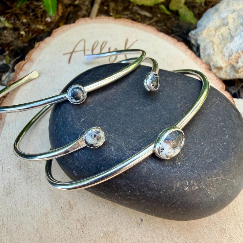 May include: Three silver-toned, open-ended bangles with a small, round, speckled stone at each end. The bangles are resting on a black stone and a wooden surface. The word "Allyk" is written on the wooden surface.
