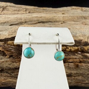 May include: Silver-tone earrings featuring round, turquoise-coloured stones. The earrings are presented on a white stand, set against a wood-grain backdrop. The turquoise stones have a marbled appearance.