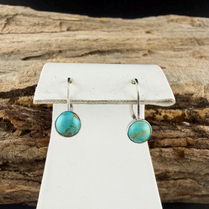 May include: Silver-tone earrings featuring round, turquoise-coloured stones. The earrings are presented on a white stand, set against a wood-grain backdrop. The turquoise stones have a marbled appearance.