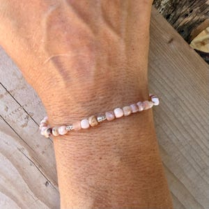 May include: A delicate beaded bracelet featuring small, smooth, light pink and peach-colored stone beads interspersed with tiny, cylindrical silver-toned metal spacers. The beads are uniform in size and arranged in a single strand around the wrist.