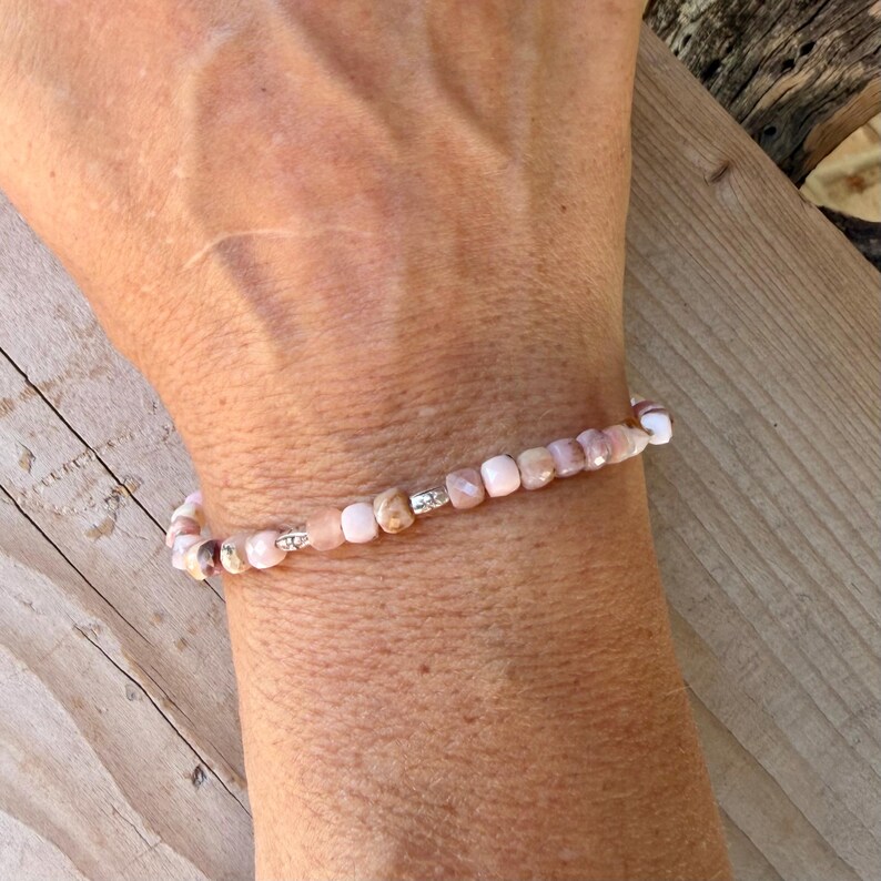 May include: A delicate beaded bracelet featuring small, smooth, light pink and peach-colored stone beads interspersed with tiny, cylindrical silver-toned metal spacers. The beads are uniform in size and arranged in a single strand around the wrist.